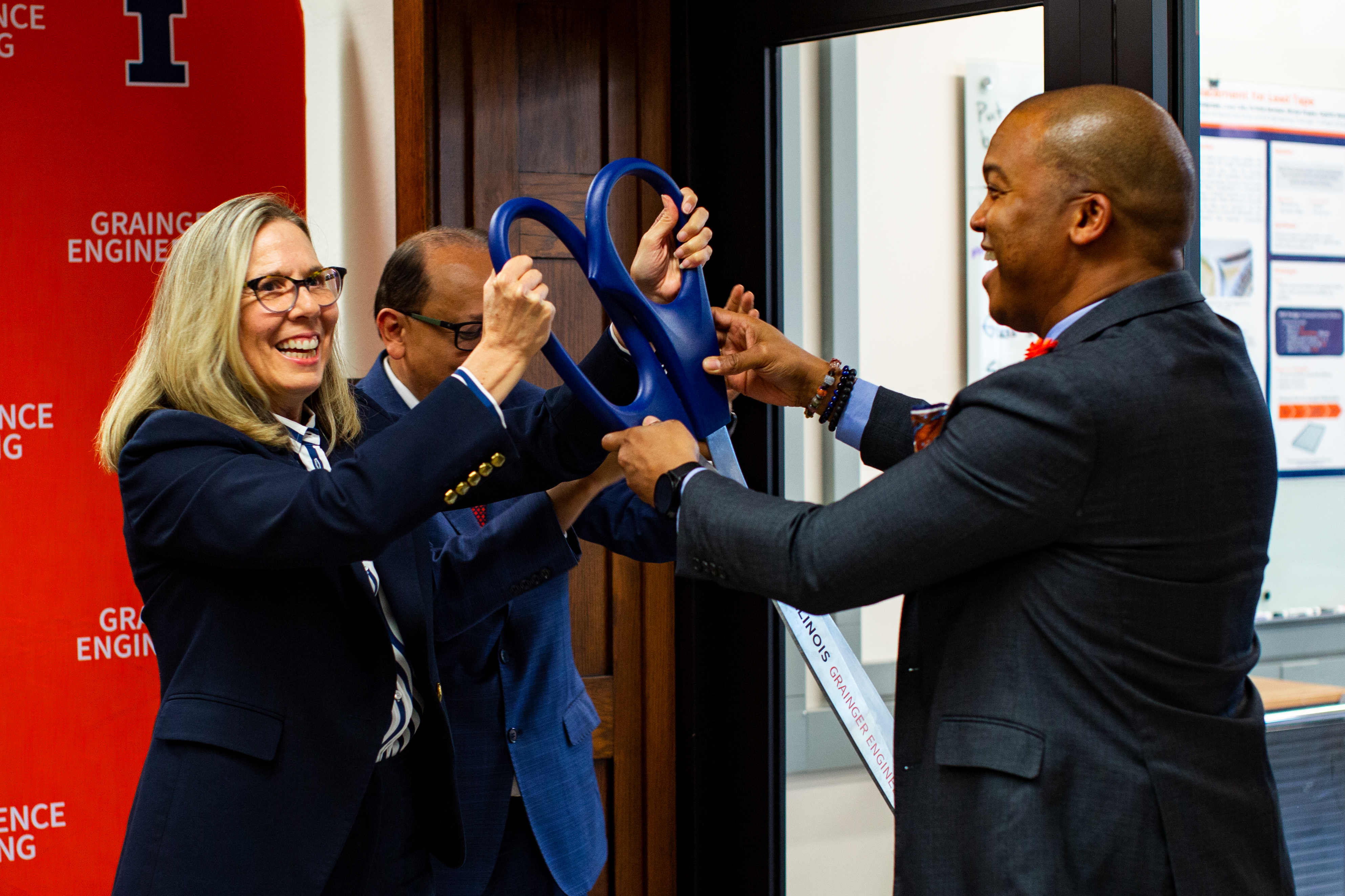 Nancy Sottos, Rashid Bashir and Dale Wright smile as they hold a giant pair of scissors moments before the ribbon-cutting happens.