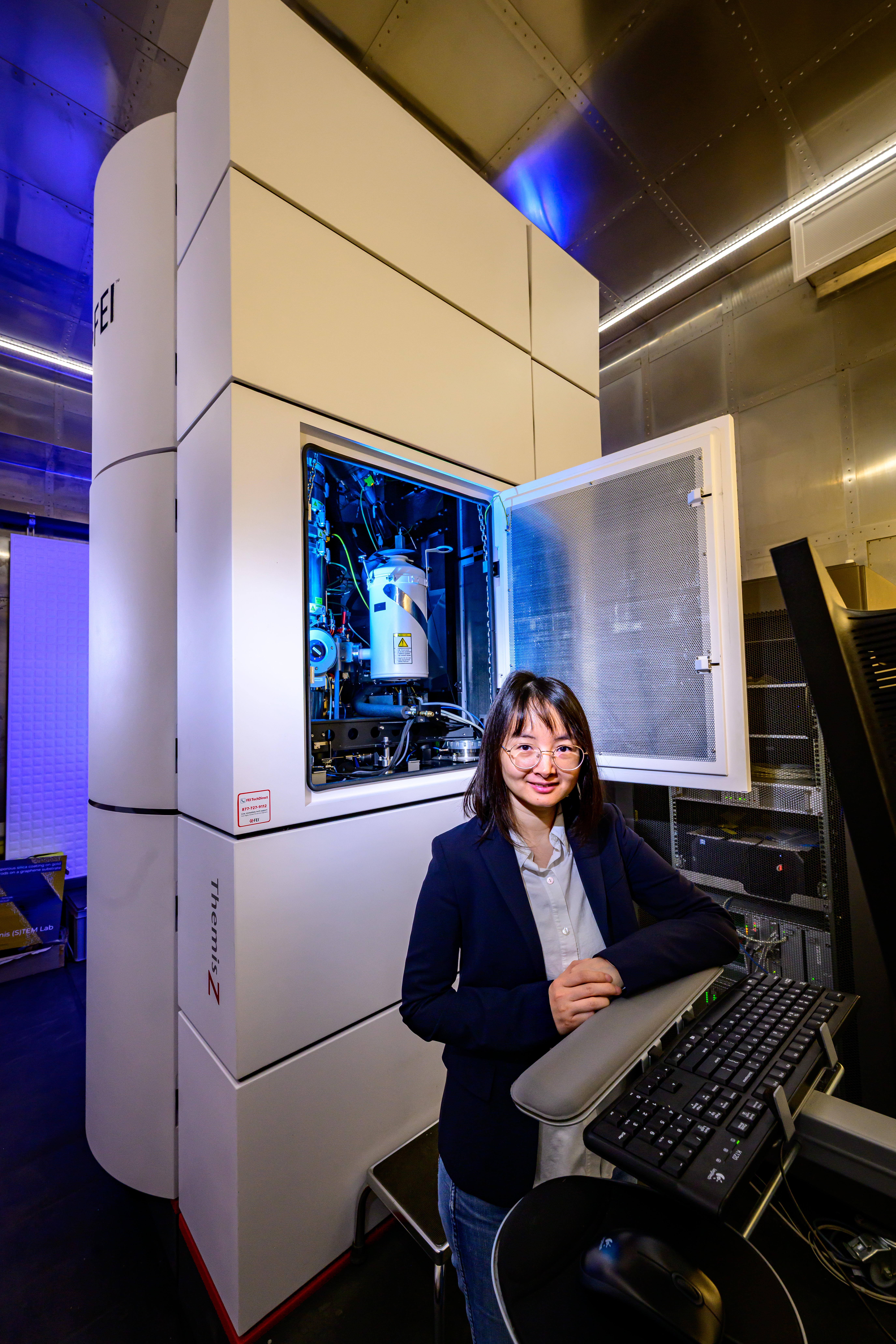 Qian Chen stands in front of microscopy equipment and smiles