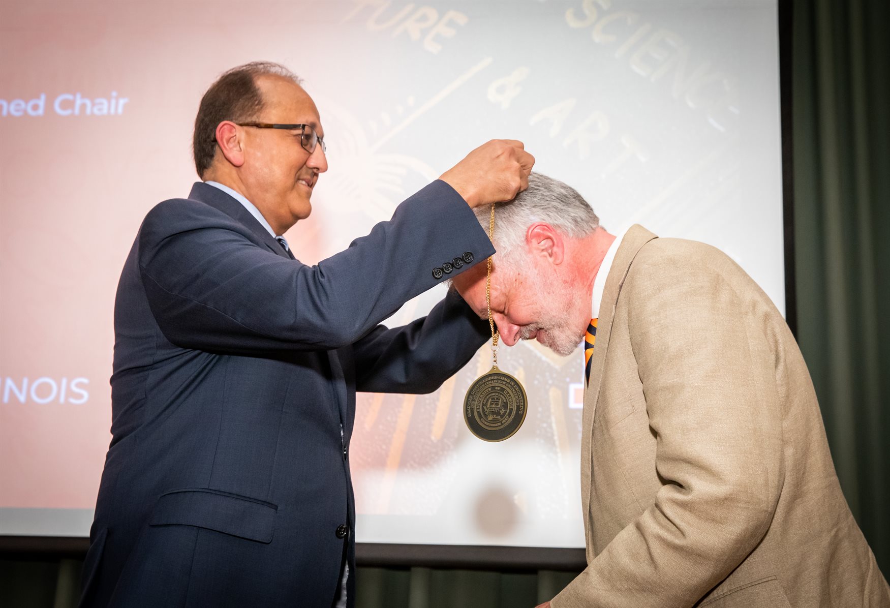 Fred Zwicky/Illinois News Bureau<br>Dean Rashid Bashir, left, bestows David Cahill his medallion for his investiture as the Grainger Distinguished Chair in Engineering during the Engineering Investiture Ceremony at the Beckman Institute in Urbana, Ill. on Monday, May 2.<br><br>Bashir shared that like Paul Braun, he, too, had to whip out his Macbook to feverishly take notes while in a meeting with Cahill about one of his research efforts.<br><br>"I was trying to use silicon transistors to do surface heating in fluids. I pretty much had to go back and rewrite the paper after the meeting," Bashir joked.<br><br>Jokes aside, the Grainger Engineering dean said he admires Cahill's leadership, especially when he was MatSE's department head.<br><br>"One of my aspirations (has) always been to do administration but then make it integrated with research so essentially stay research active," Bashir said."I used to look at David as a department head (and see) that he continued to do so, (and) I really, really appreciate that."